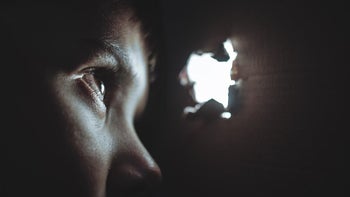 A woman is looking out through a hole in a wall spying on people.