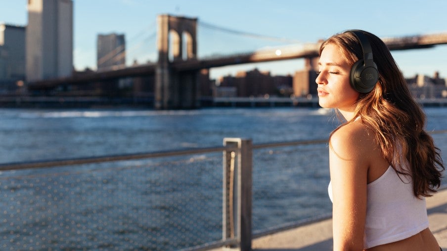 A woman wearing JBL Tune 760NC headphones enjoys the sunlight by a waterfront with a city skyline and bridge in the background