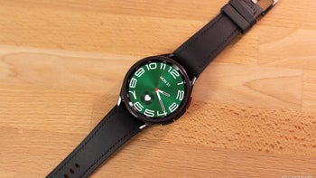 A Galaxy Watch 6 Classic with a black leather band displaying a green dial with large white numbers, lying flat on a wooden table.