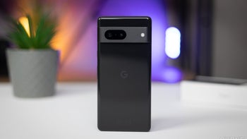 A Google Pixel 7 stands upright, showcasing its black glass back panel, against a background of soft purple and orange lighting with a blurred potted plant and circular light reflections.