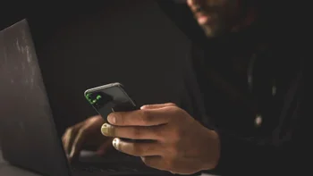 Scammer in the dark holding an iPhone and looking up something on his laptop.