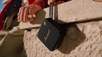 A Marshall Willen portable speaker hangs by its strap on a stone wall. There's a hand nearby.