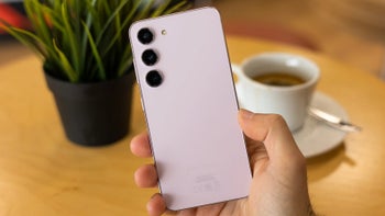 A person's hand holding a pink Samsung Galaxy S23 smartphone above a wooden table with a potted plant and a coffee cup.