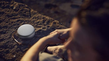 A person sitting on a beach with a Bang & Olufsen Beosound A1 (2nd-gen) speaker nearby