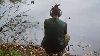 A man standing in front of a river with headphones on his head, his back facing the camera.