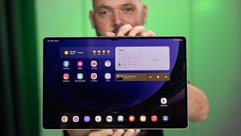 A person holding the Galaxy Tab S9 Ultra with a turned-on display, set against a blurred green background.