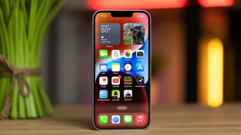 An iPhone 14 on a wooden table, displaying the home screen with various app icons and widgets.