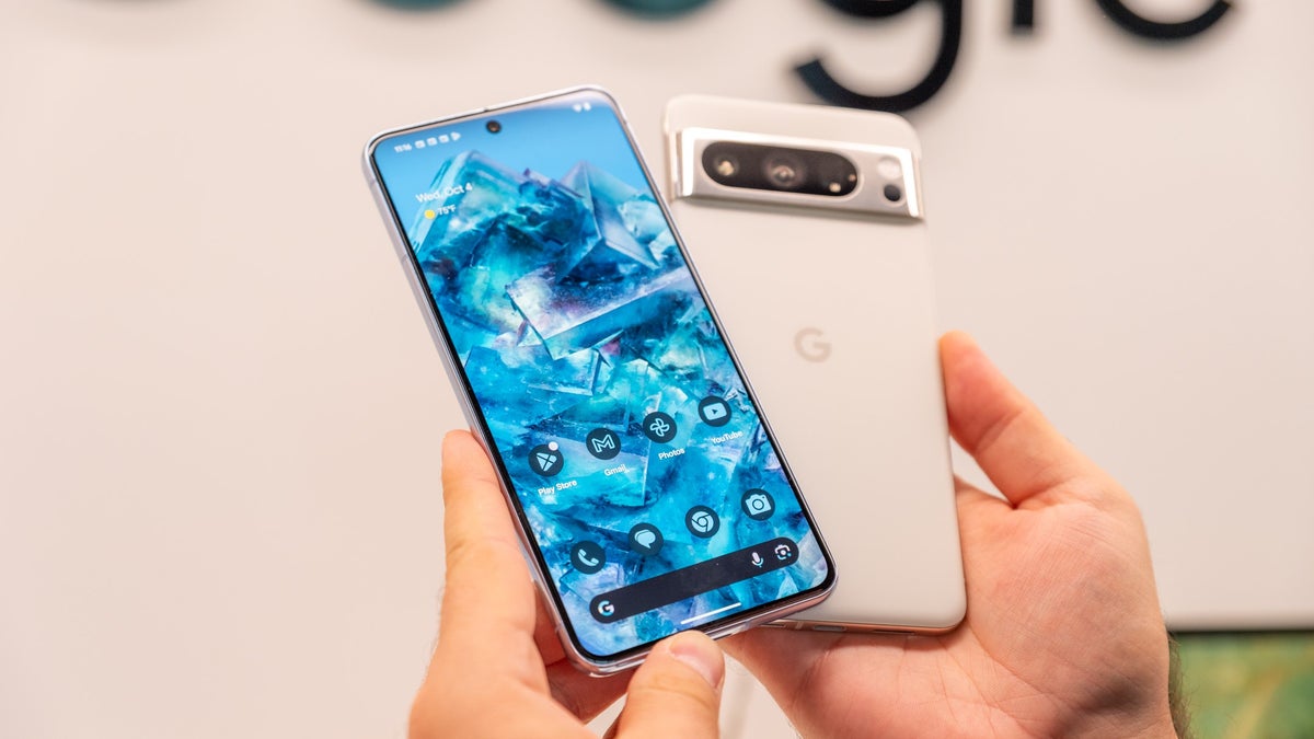 Hands holding the Pixel 8 Pro, showing the front display with app icons and the rear camera module.