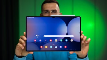 A person with eye glasses holding the Galaxy Tab S10 Ultra against a blurred green background, showing its home screen.