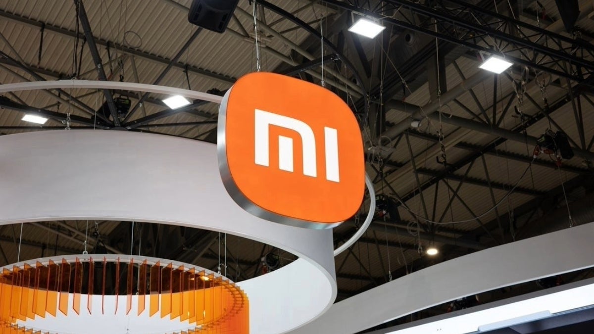 The Xiaomi logo, a white "Mi" in a square orange box, hanging from the ceiling of a large, brightly lit room.