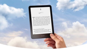 A person's hand holding a black Kindle e-reader against a blue sky with white clouds.