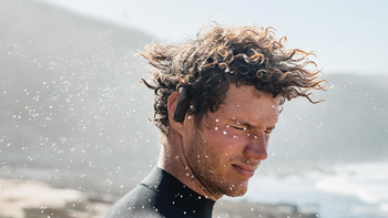 Man with curly hair wearing JBL Endurance Peak 3 workout earbuds, standing near the sea with water splashing around him.