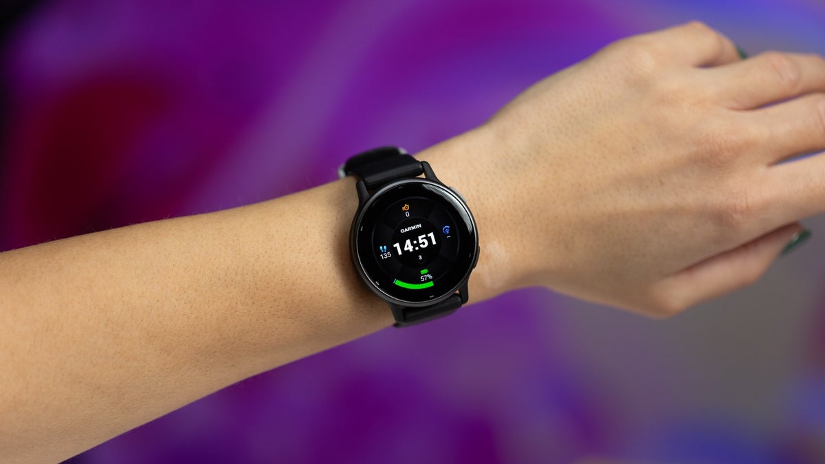 A person's arm wears a Garmin Vivoactive 5 smartwatch, showcasing its display against a blurred purple background.