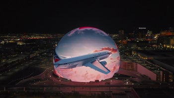 A large, spherical building is illuminated with a dynamic projection of a Delta Airlines airplane flying through a sunset sky.