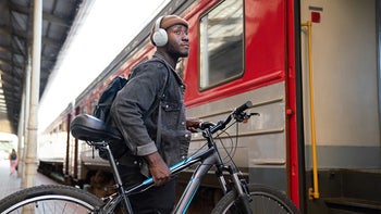 A person with a backpack and JBL Tune 770NC headphones holding a bicycle while standing at the entrance of a train, ready to board.