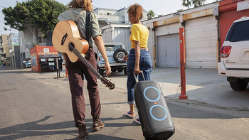 Two people walk down an alley, one carrying an acoustic guitar on their back and the other pulling a PartyBox 310 speaker on wheels.