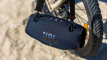 The JBL Xtreme 4 portable Bluetooth speaker hangs by its strap on a bike in a sandy outdoor setting.