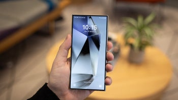 A close-up of a hand holding the Galaxy S24 Ultra against a wooden table showcasing its display.