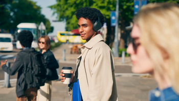 A young man wearing Sony WH-CH720N headphones, holding a coffee cup, and smiling while walking in an urban setting.