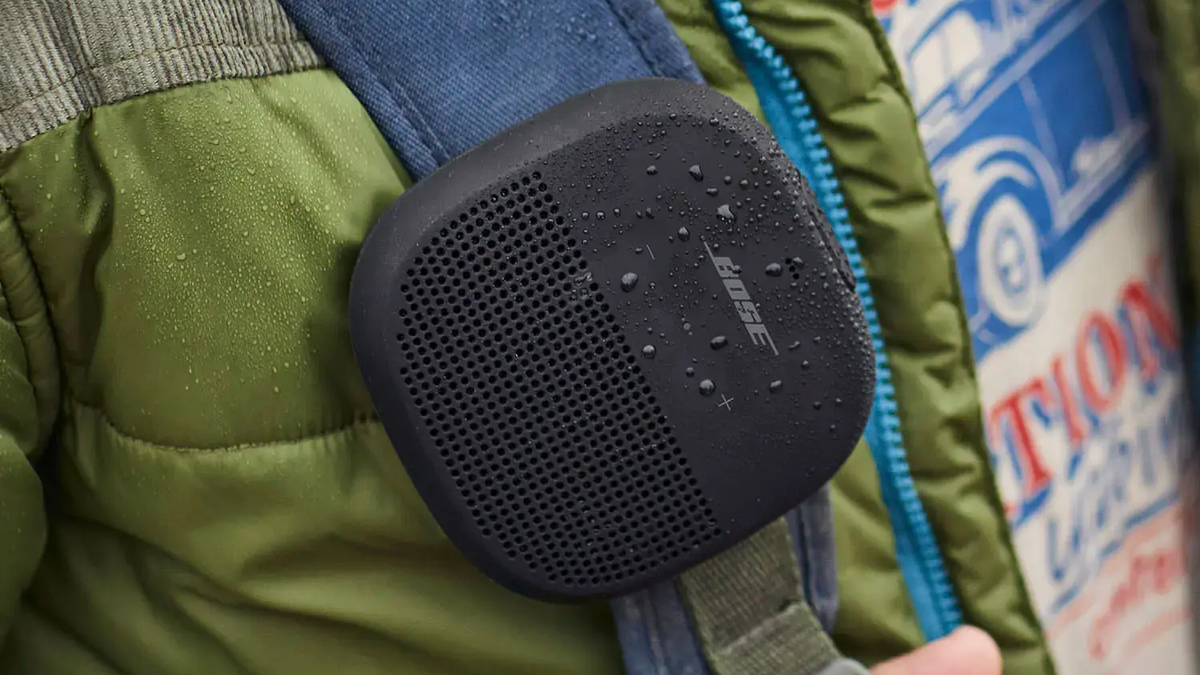 A Bose SoundLink Micro speaker with water splashes is attached to a backpack, highlighting its waterproof design.