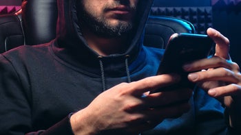 A scammer dressed in a hoodie checks out how his latest scam is doing on his phone.