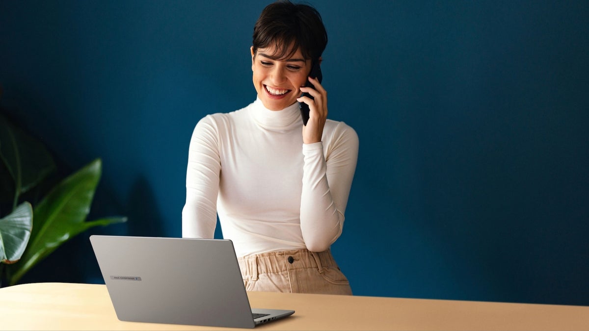 A photo of a person smiling while talking on a smartphone and looking at a laptop on a desk.