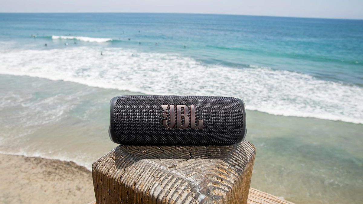 A JBL Flip 6 speaker near the sea, with sand and waves visible in the background.