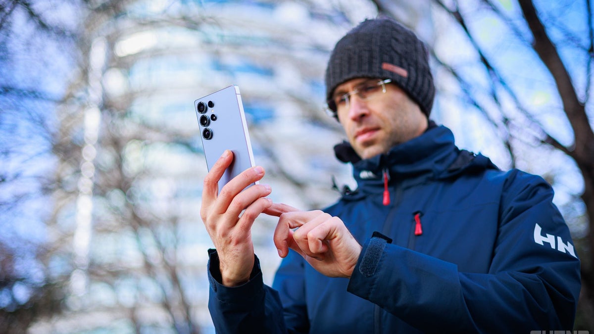 Man dressed in winter clothes holding a Galaxy S25 Ultra and using it.
