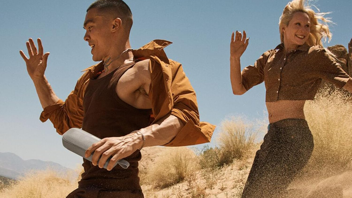 A smiling man is running downhill on sand while holding the Bose SoundLink Flex 2nd Gen in Sandstone.