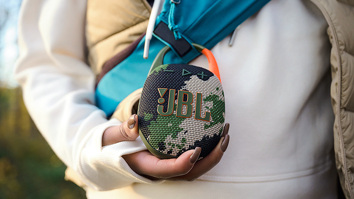 JBL Clip 5 is clipped on a backpack with its iconic logo visible on the speaker's front side.