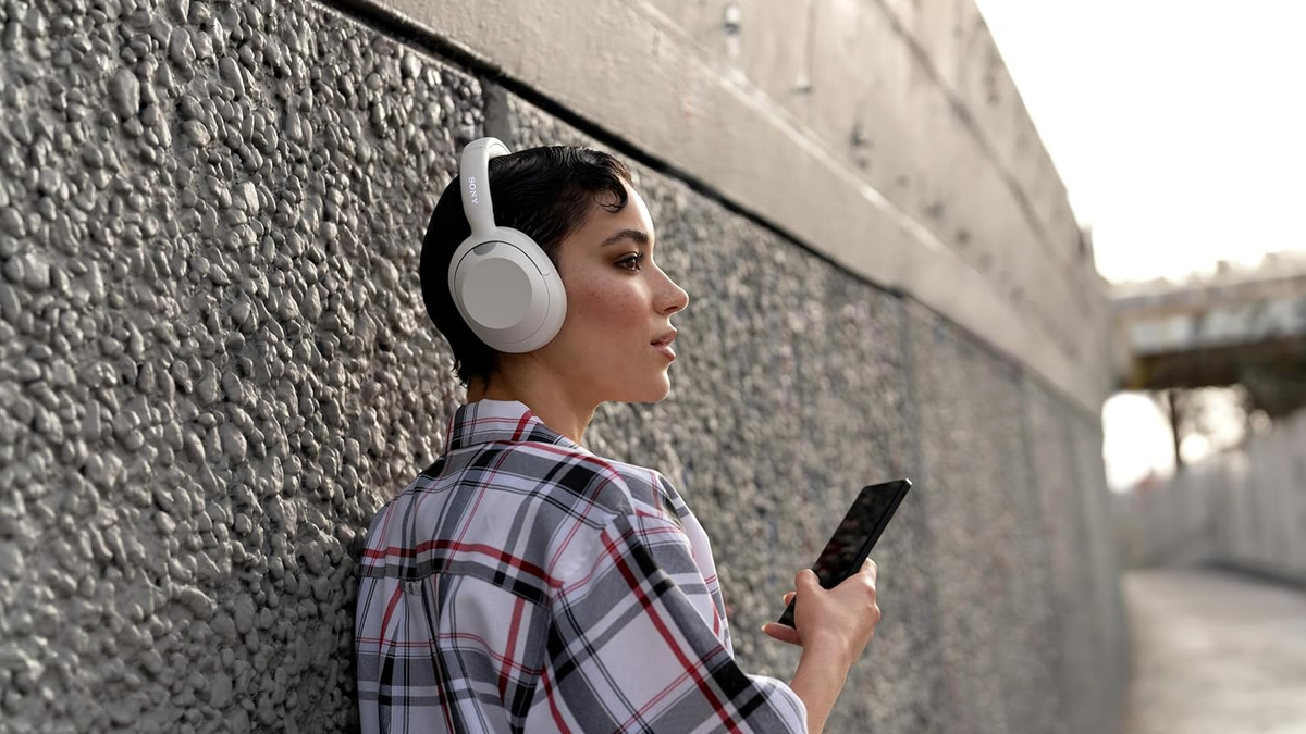 A woman leaning on a wall is wearing the Sony ULT Wear headphones while holding her smartphone.