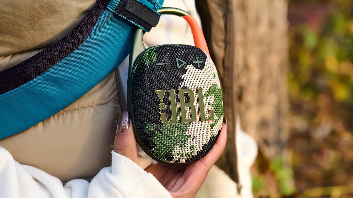 A close-up of a JBL Clip 5 speaker attached to a person's backpack.