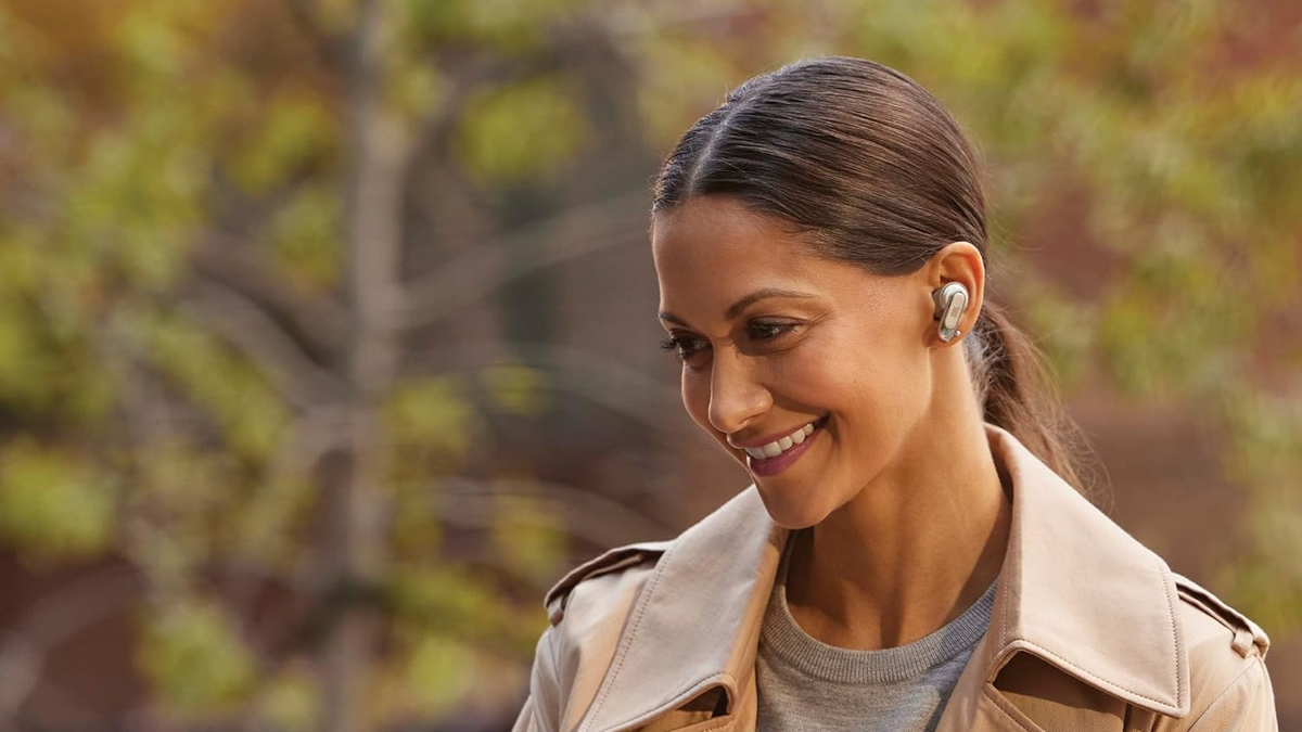 A smiling woman wears the JBL Tour Pro 2 earbuds in an outdoor setting, with trees visible in the background.