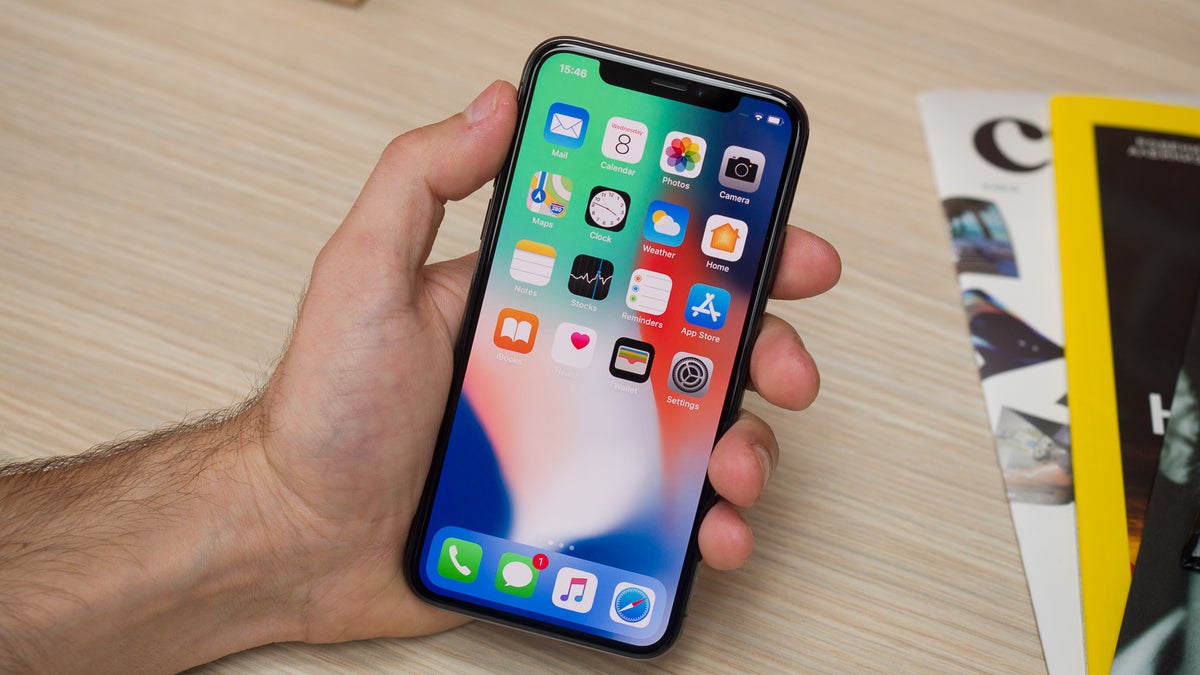 An iPhone X in a male hand with its display showing the home screen.