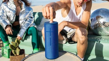 Two people sitting outside, one is pressing a button on a portable speaker.