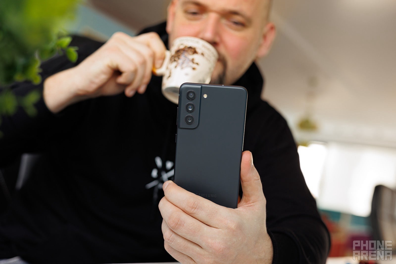 A person drinking coffee in the background while holding a Samsung Galaxy S21 FE device