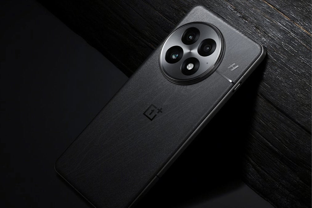 A black OnePlus 13 smartphone is shown resting on a wooden surface.