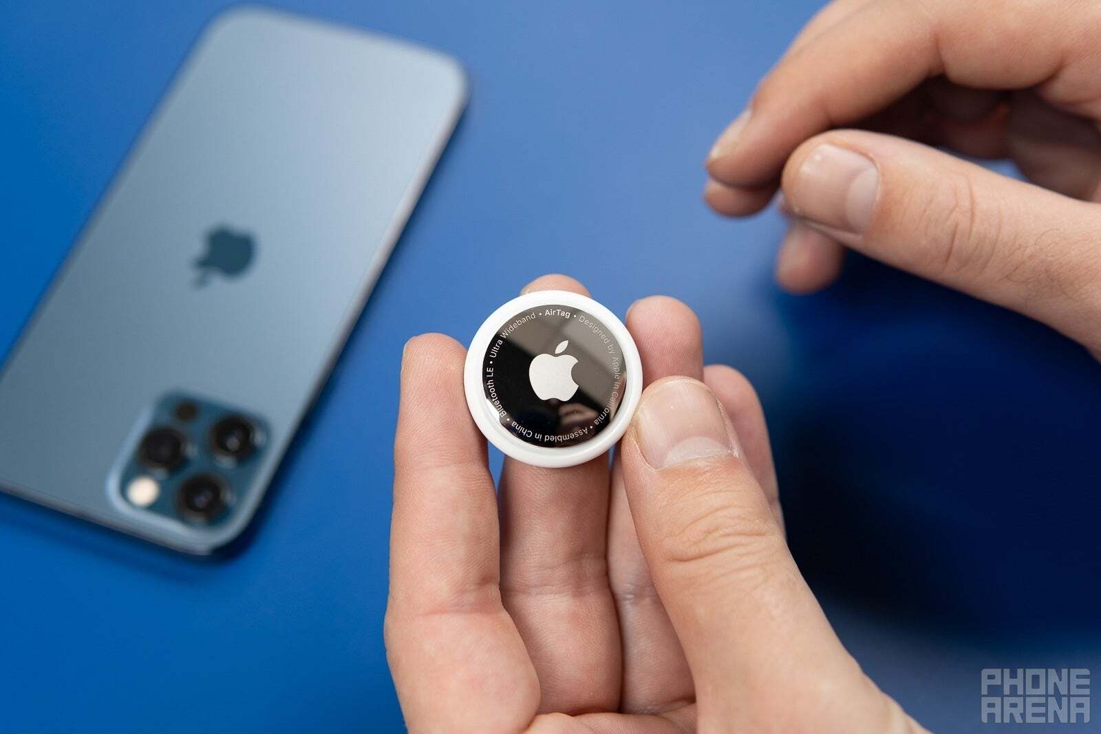 An Apple AirTag held by someone&amp;#039;s hand with an iPhone in the background