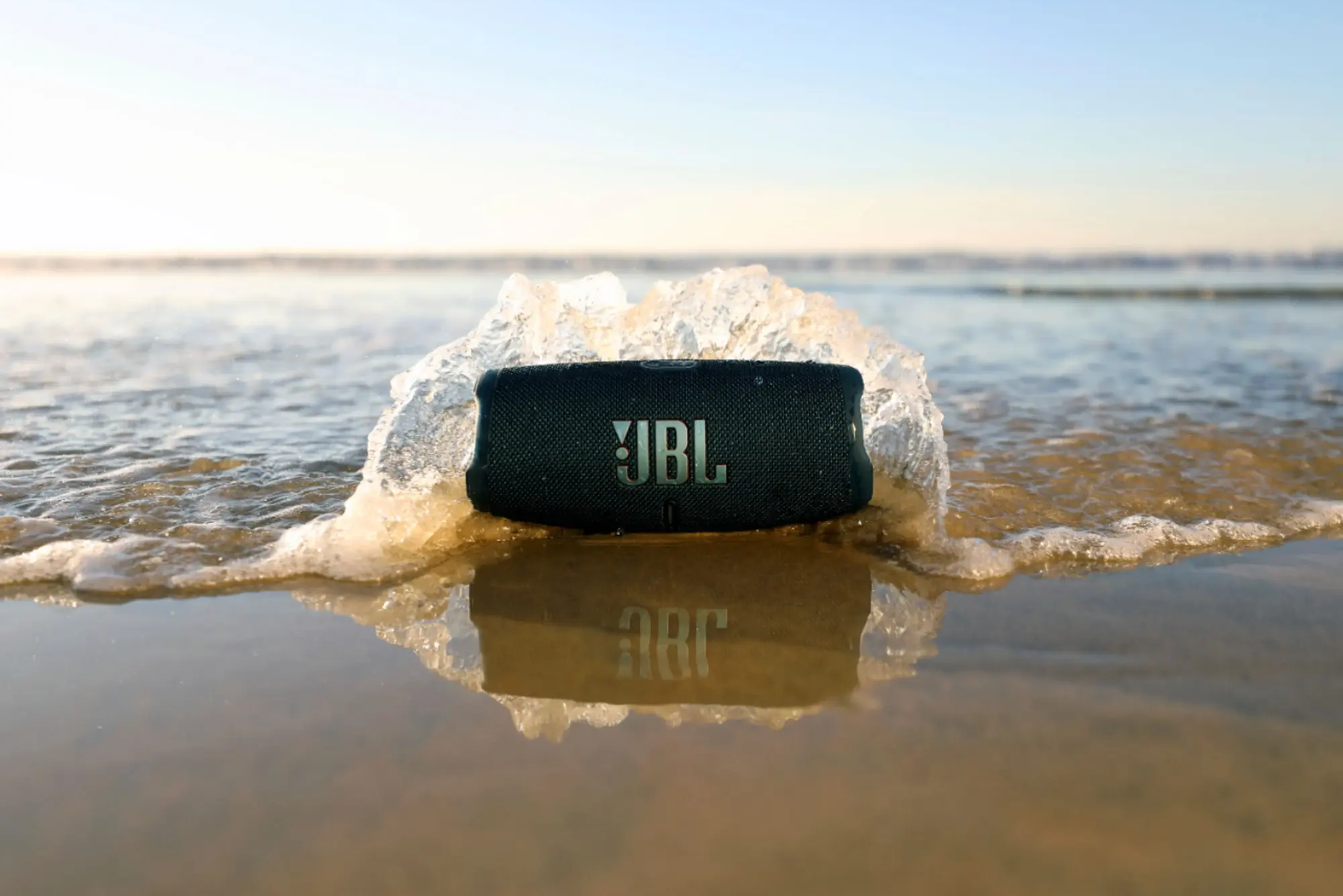 A JBL Charge 5 speaker sits on a beach as ocean waves splash around it, reflecting on wet sand.