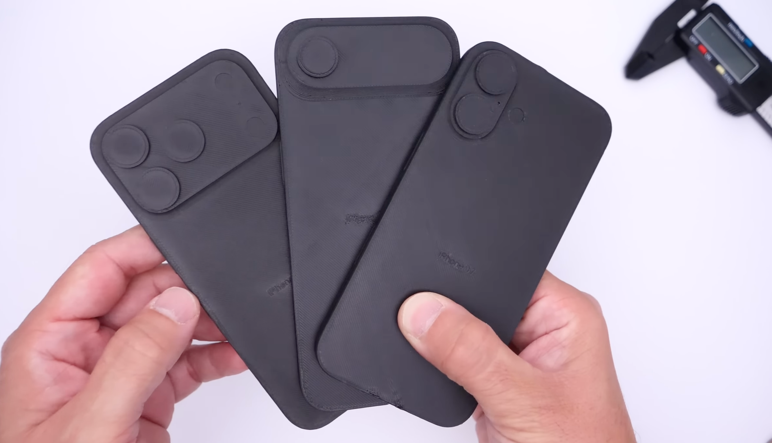Two hands holding three black iPhone dummies with different camera cutouts.