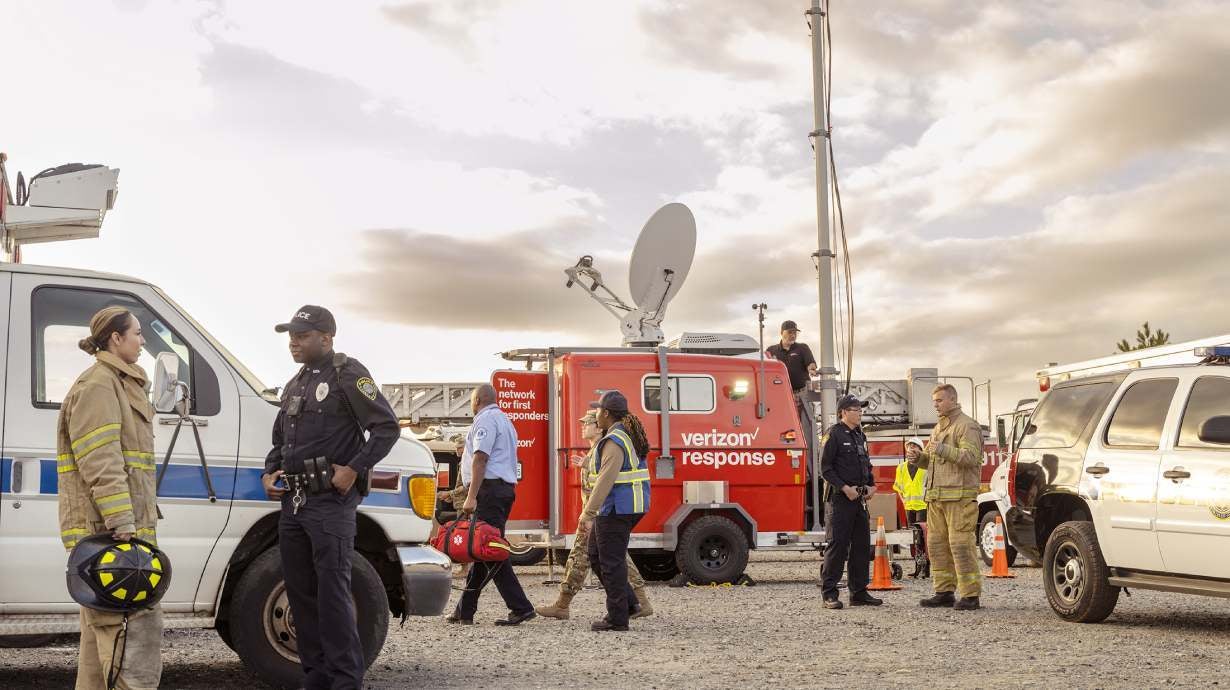 Several emergency responders, including firefighters and police officers, are gathered around a Verizon Response vehicle equipped with a satellite dish.