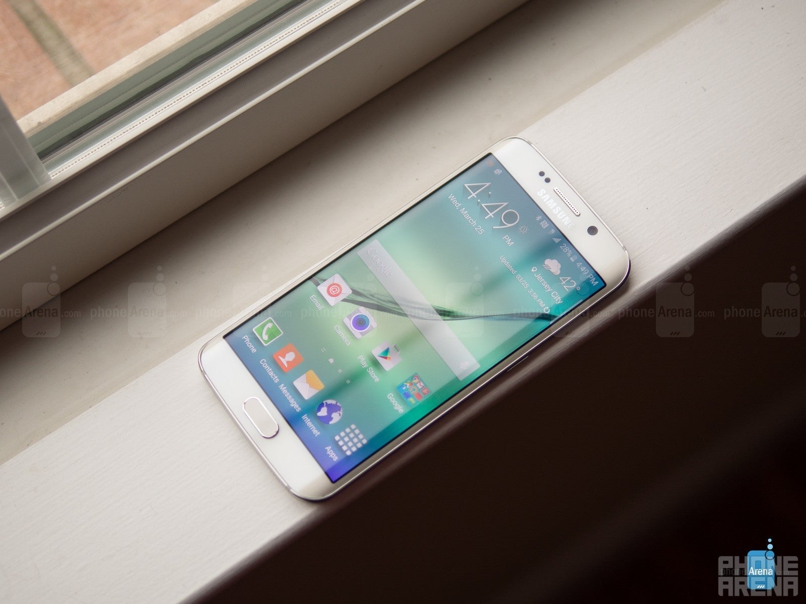 Samsung Galaxy S6 Edge em uma janela, retratada diretamente de cima. A tela está ligada, mostrando a tela inicial