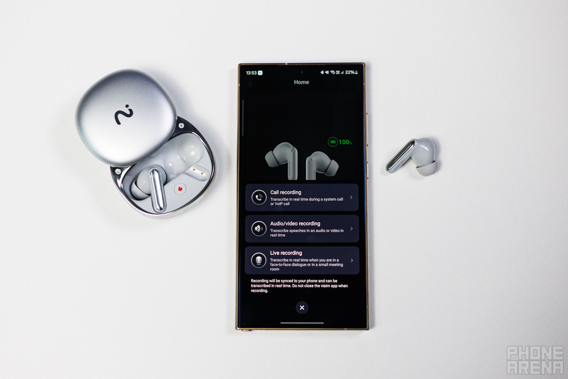 Viaim RecDot placed on a table with the flap of its case open. The right earbud is outside the case, and its laying next to a Samsung Galaxy S24 Ultra, showing the Viaim app on screen