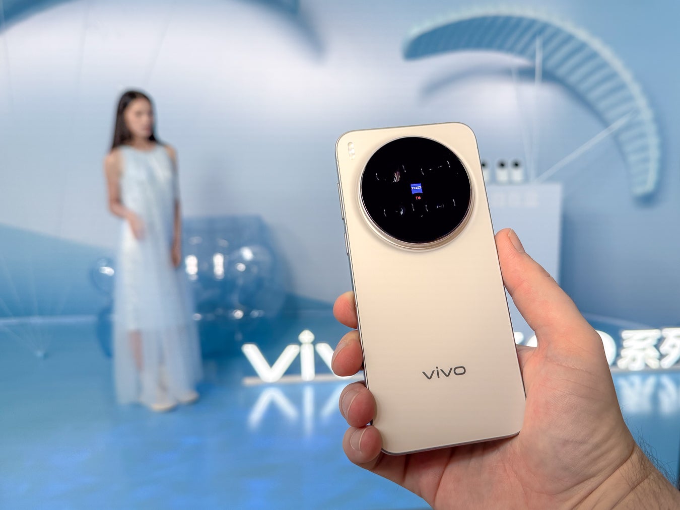 Person holding a Vivo X300 Pro in gold; its large circular Zeiss-branded camera module visible; a model posing in the blurred background at a Vivo event.