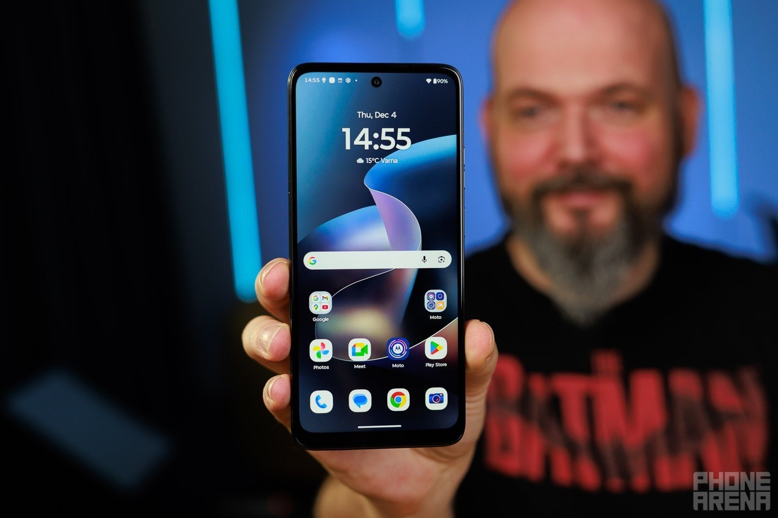 Person holding a Motorola smartphone facing the camera, with the home screen visible against a blurred studio background.
