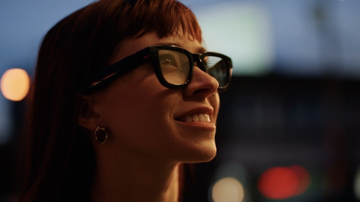 Person wearing smart glasses at night, smiling with city lights blurred in the background.