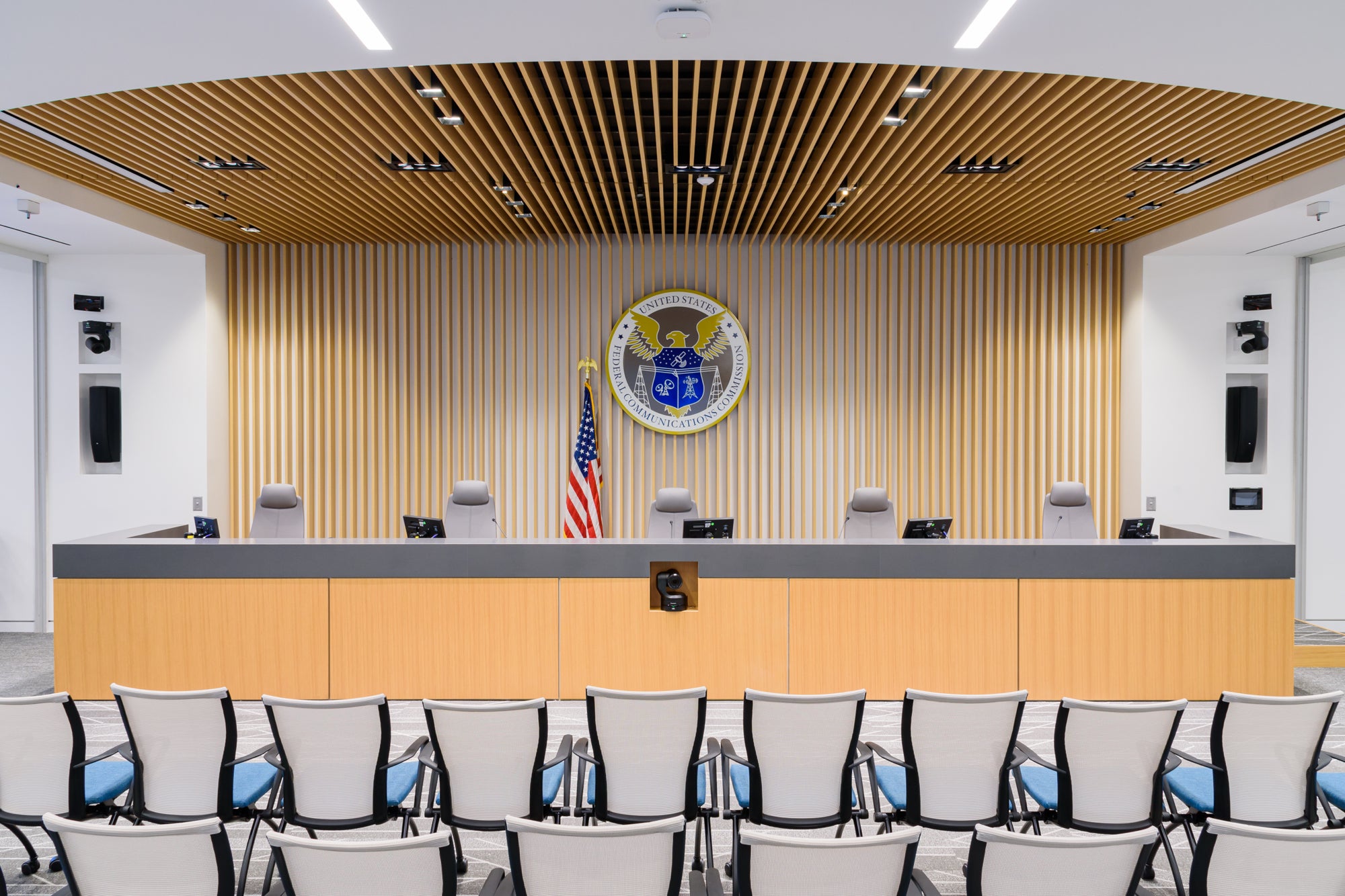 The dais and five commissioner seats inside the FCC&amp;#039;s commission meeting room, with a United States flag and the FCC seal on the wall behind