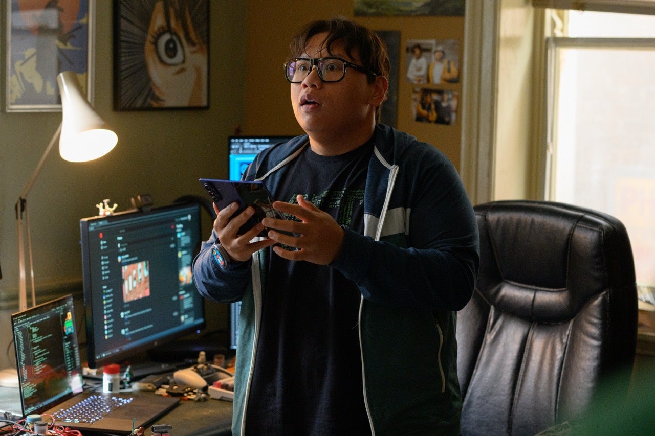 Ned Leeds, played by Jacob Batalon, reacts while holding a Samsung Galaxy phone at a desk covered in monitors.