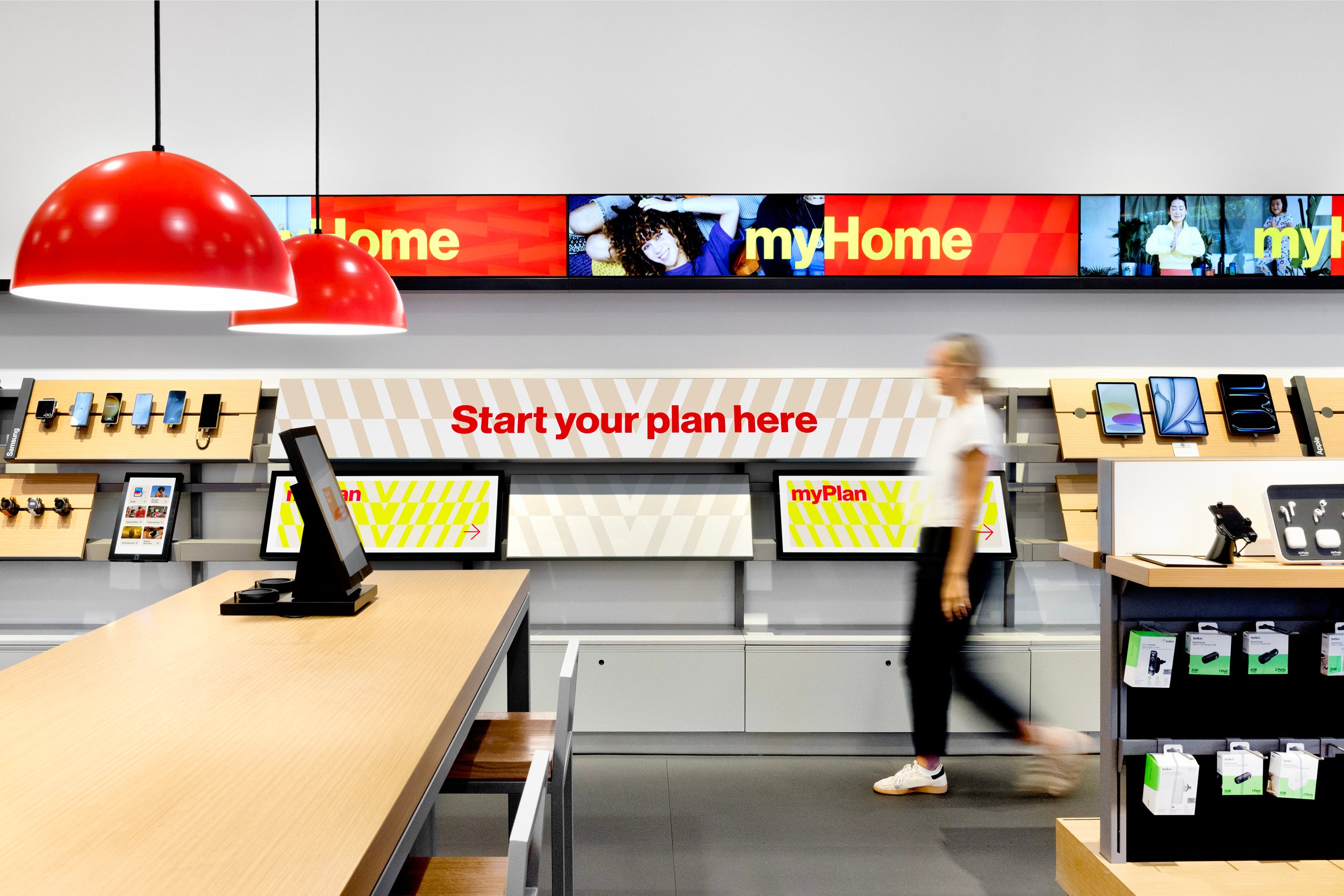 nterior of a Verizon retail store with smartphone and accessory displays along the walls, red dome pendant lamps overhead, myPlan and Start your plan here signage, and a person mid-stride across the sales floor.