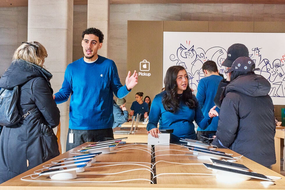 Apple Store team members in blue shirts talking with customers on the sales floor.